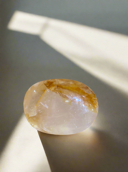 An oval-shaped Golden Healer Quartz crystal with a polished surface, placed on a shadowed surface, with a warm golden glow.