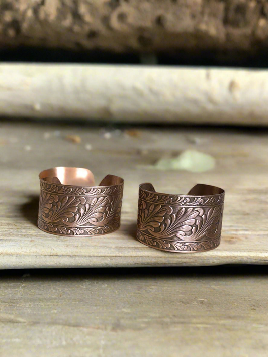 Two engraved copper cuff bracelets with a floral design on a wooden surface.